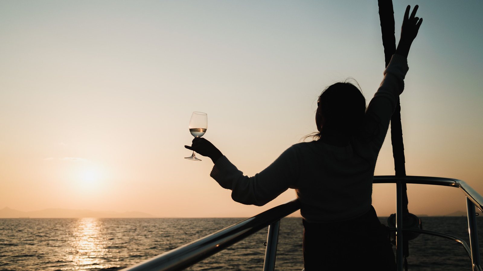 Silhouette of a person holding a glass of wine at the rail of a boat at sunset