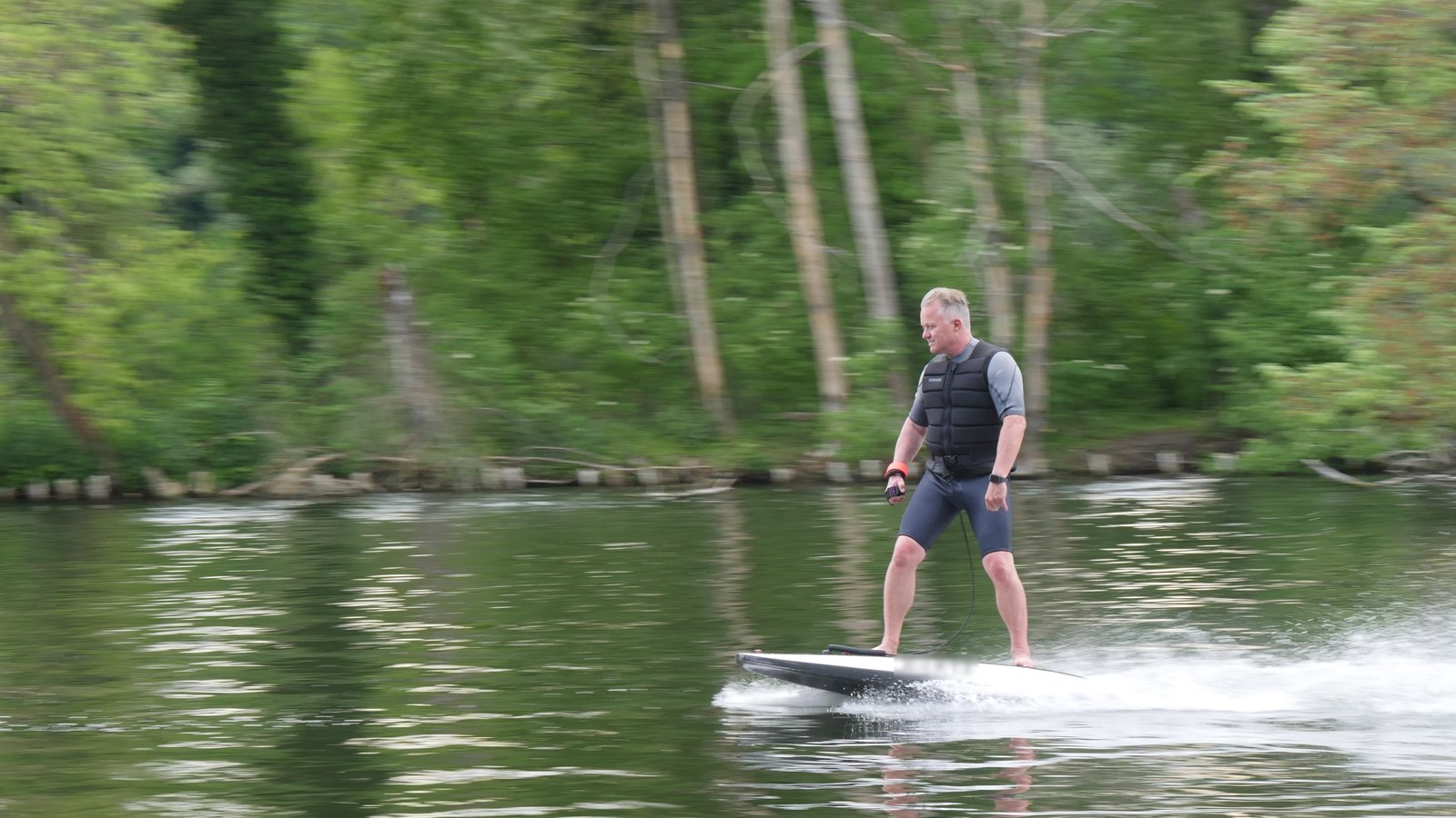 A rider in a wetsuit and impact vest gliding fast on a white electric jetboard past a wooded shore