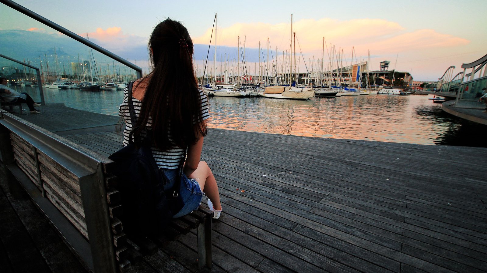 Young woman with a backpack sitting on a bench, looking out over a marina at sunset