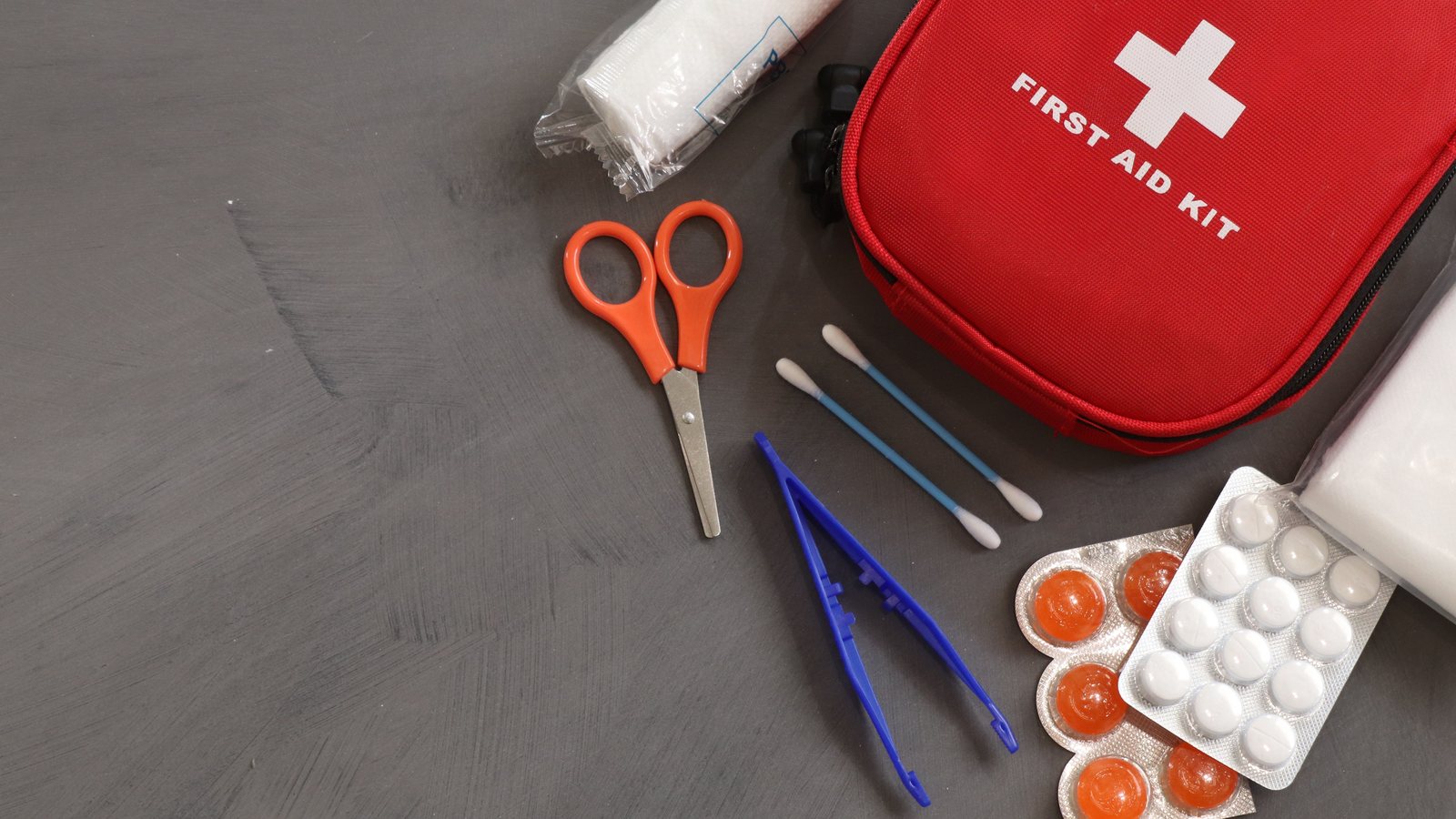 Red first aid kit with bandages, scissors, tweezers and tablets on a grey surface