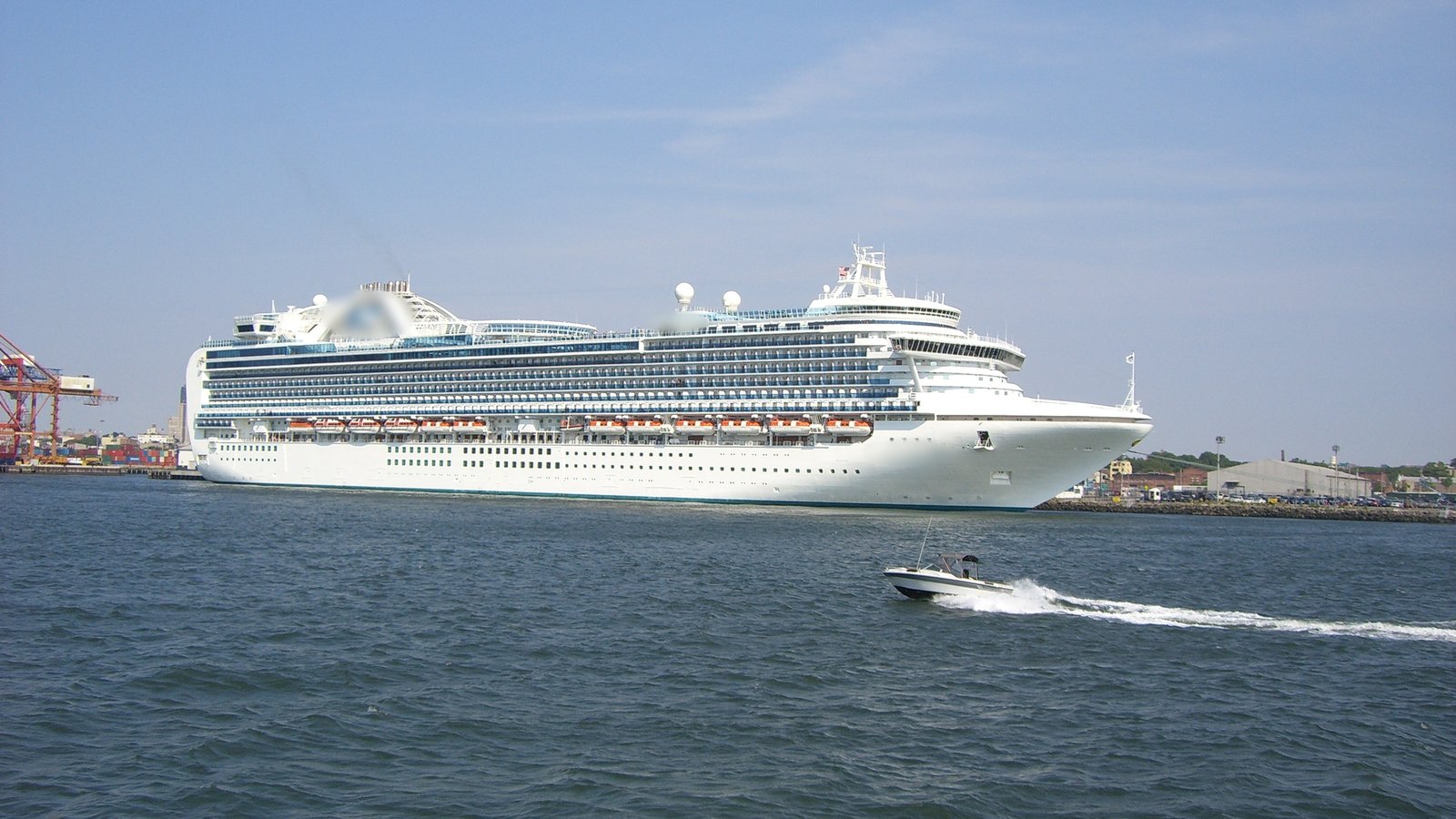 A large cruise ship in port with a small motor boat speeding past in front