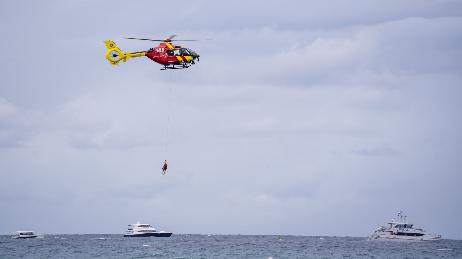 Rescue helicopter winching a person above the sea, a motor yacht and a police boat below