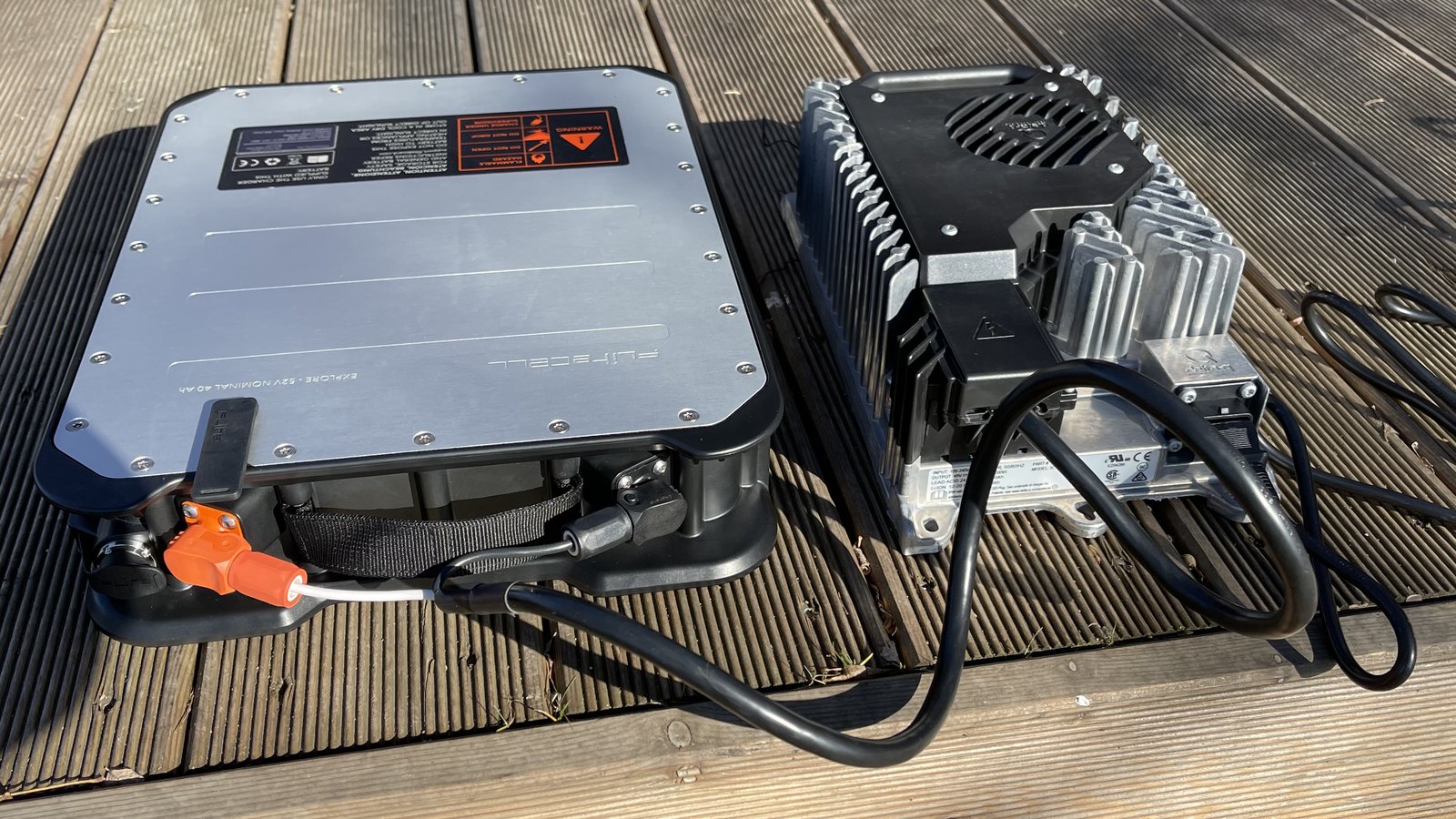 A flat aluminium eFoil battery pack cabled to a finned charger on a wooden deck