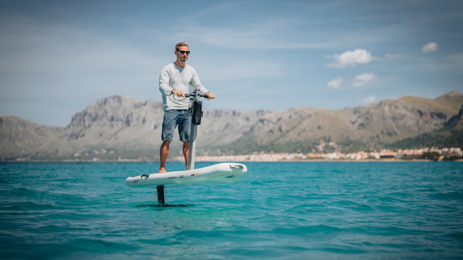 A rider standing at the handlebar of an eFoil scooter, flying above turquoise water with mountains behind