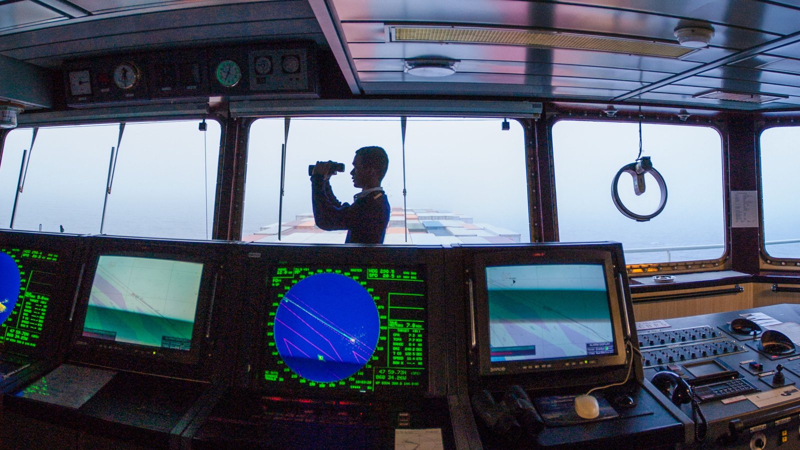 Officer with binoculars on the bridge of a container ship, radar screens in the foreground