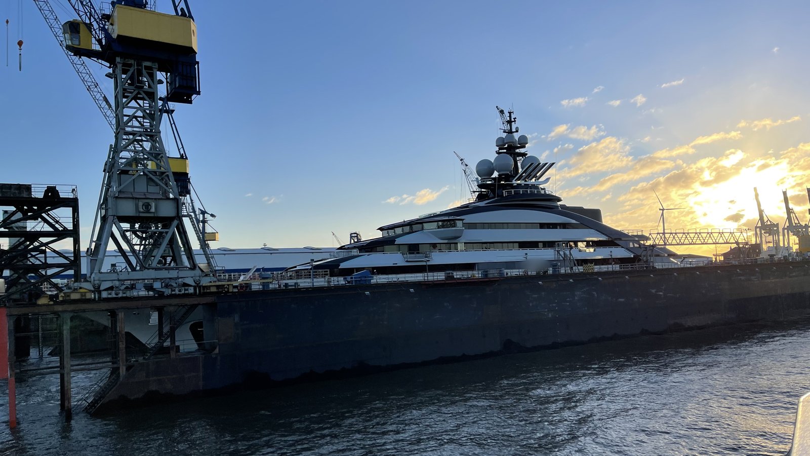 A large superyacht in a floating dock between shipyard cranes, evening sun behind