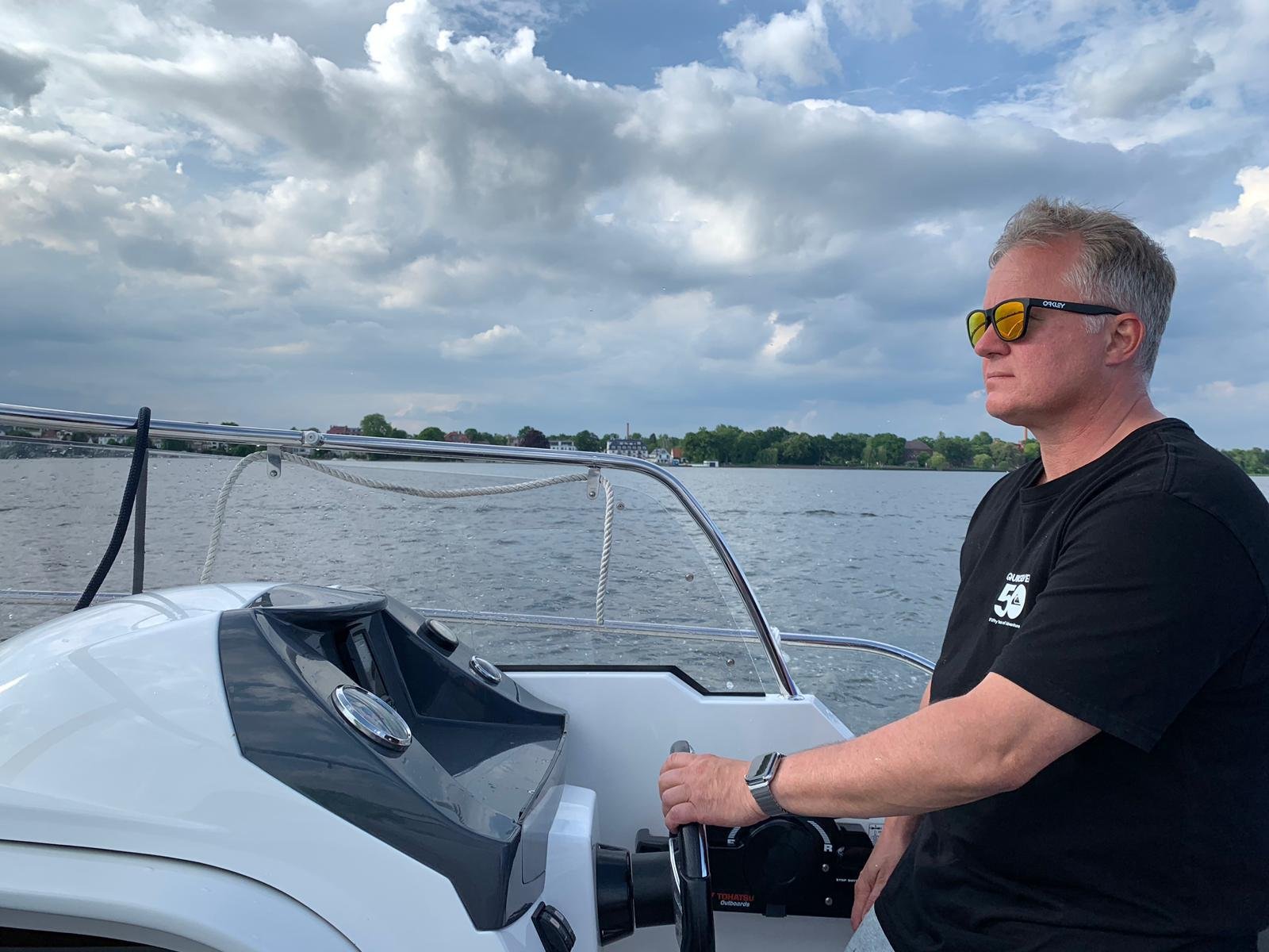 Andreas Lakeberg in sunglasses at the helm of a small motor boat on a lake under a cloudy sky